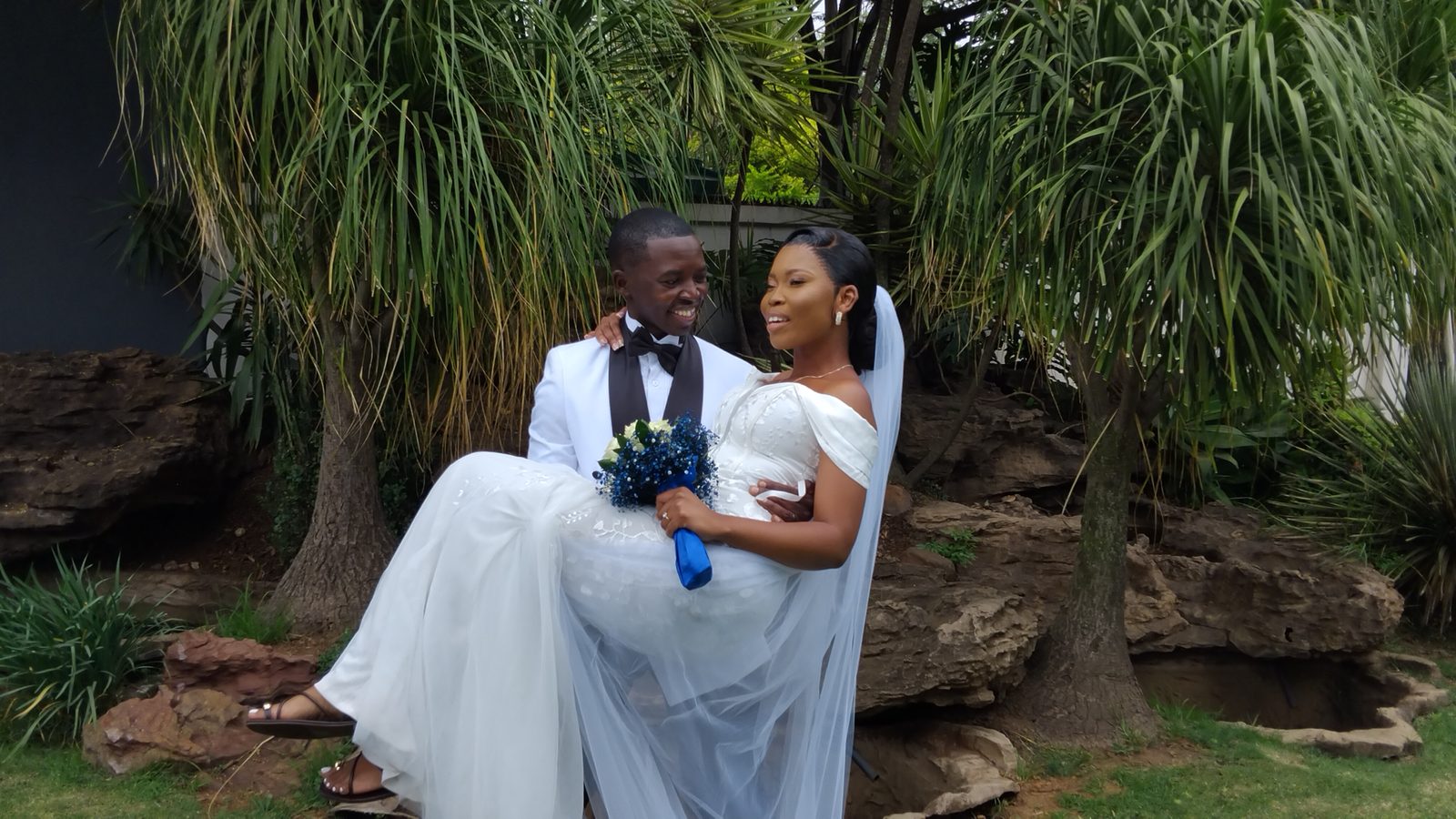 A groom in a white jacket carrying a smiling bride in a garden.